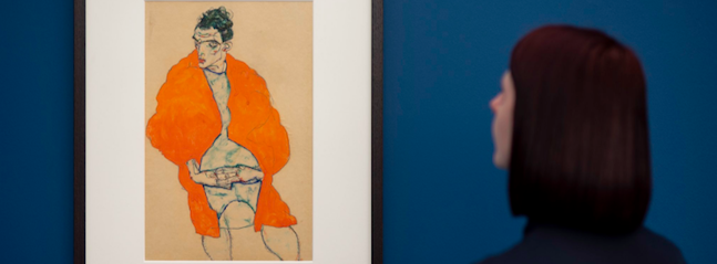 A person with shoulder-length dark hair and a dark shirt is viewed from behind, standing and looking at a framed painting of a figure wearing a bright orange coat on a blue and beige background. The scene is set against a dark blue wall.