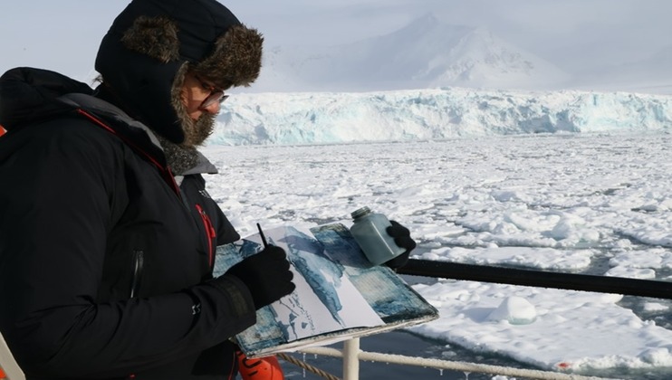 A person bundled in winter clothing stands on a ship, painting in a sketchbook. They are surrounded by an icy, snow-covered landscape with a distant mountain, icebergs, and frozen water. The person is holding a brush and a container that appears to hold paint.