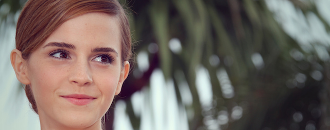 A woman with light skin and short, side-parted brown hair is smiling softly while looking to the side. She is outdoors with a blurred background featuring palm tree fronds.