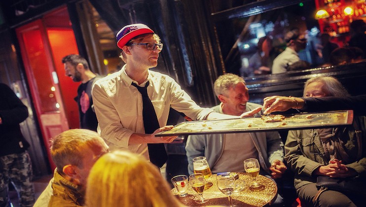 A man wearing a white shirt, black tie, and red hat serves food on a tray to a group of people at a lively bar. There are drinks on the table, and the bar is warmly lit with a bustling atmosphere. Other patrons can be seen in the background enjoying the evening.