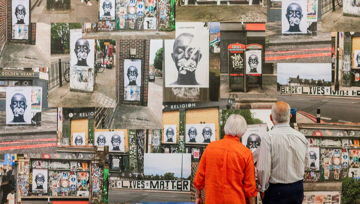 Two elderly people stand before an urban collage wall adorned with numerous pasted images and posters. Prominent amongst the images is a graphic face with flowers covering the mouth. Visible posters include BLACK LIVES MATTER and various graffiti.