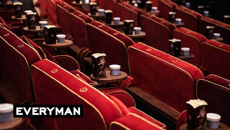 A theater with rows of plush, red velvet seats, each paired with a small wooden table holding cups and popcorn. The ambiance appears cozy and luxurious. The word EVERYMAN is displayed in white text on a black banner in the bottom left corner.