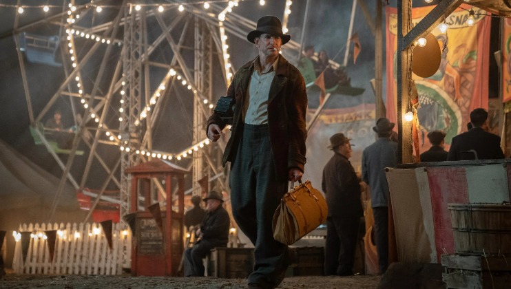 A man dressed in vintage attire and wearing a hat walks through a bustling carnival at night, carrying a yellow satchel. Behind him, a Ferris wheel is illuminated with string lights, and there are groups of people, booths, and banners in the background.