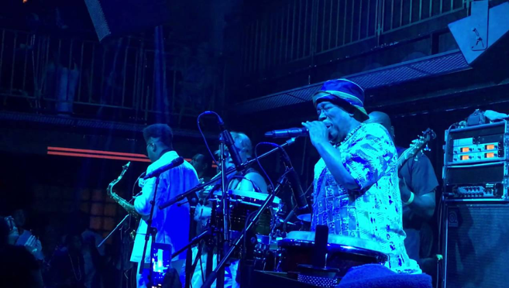 A band performs on stage under blue lighting. The lead singer passionately sings into the microphone while other band members play instruments, including saxophones and drums. An audience watches from a distance, with some individuals captured in the foreground.