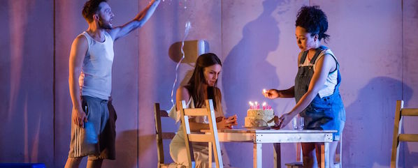 Three people are in a room. One woman sits at a table with a cake and lit candles, while another woman lights the candles. A man is standing nearby, raising his hand as if in celebration. The scene appears to depict a birthday celebration.
