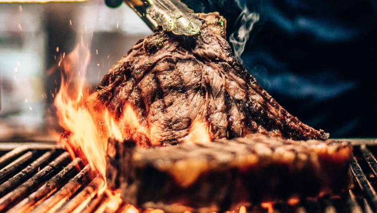 A close-up of a large piece of meat being grilled over open flames. The meat is held by tongs, showing a charred, crisp exterior evidence of high heat cooking. The ambient light highlights the sizzling and smoky surface, indicating intense grilling.