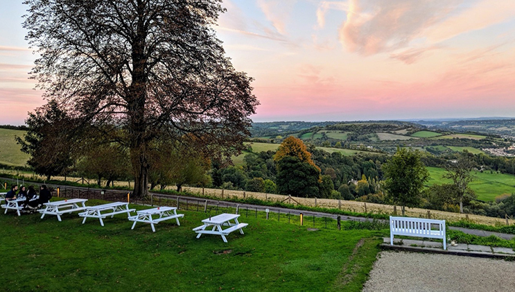 A scenic countryside view at sunset, featuring rolling green hills under a pastel sky. White picnic tables and benches are placed on the grassy area in the foreground. A large tree stands to the left, casting a shadow. The horizon shows a distant village.
