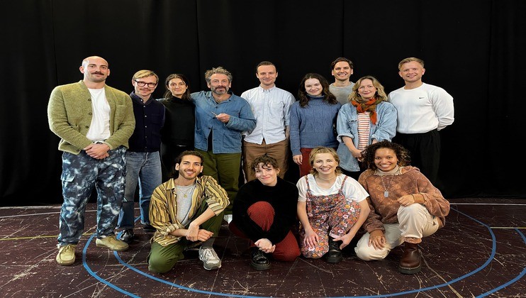 A group of thirteen people posing for a group photo indoors with a black background. They are smiling and appear to be happy. The group includes men and women, with some standing and some kneeling or sitting in front.