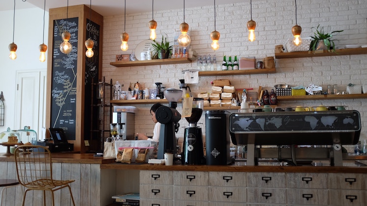 A cozy coffee shop interior with a rustic design. The counter features a chalkboard menu, a variety of espresso machines, jars, and pastries. Pendant lights hang above the wooden bar, highlighting the inviting atmosphere. Plants add a touch of greenery.