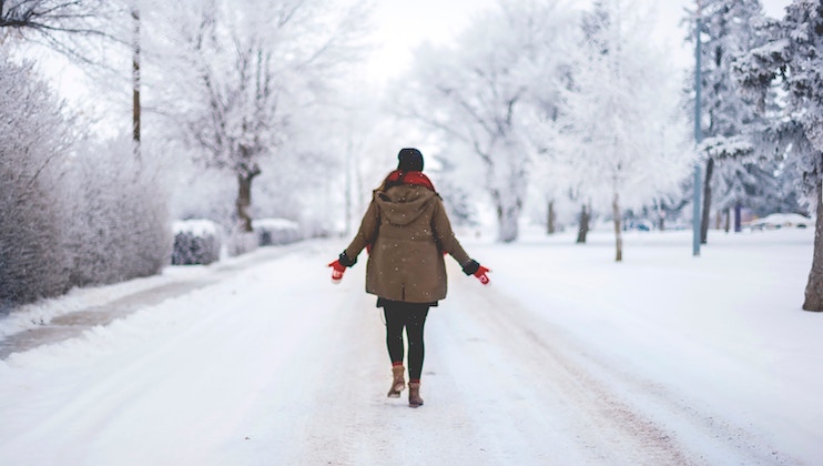 A person wearing a brown coat, red gloves, black pants, and a black hat walks on a snow-covered road lined with frosty trees. The scene is peaceful and wintery, with white snow blanketing the ground and trees.