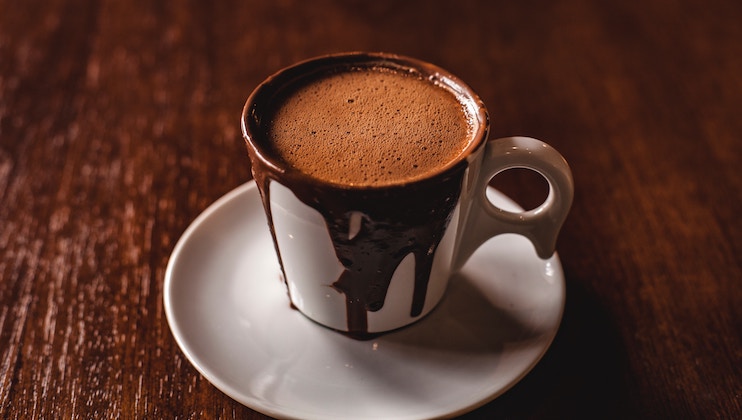 A white cup filled with a rich, frothy hot chocolate sits on a saucer. Chocolate is dripped decadently down the side of the cup onto the saucer. The background features a dark, wooden table surface, enhancing the warm and cozy atmosphere.