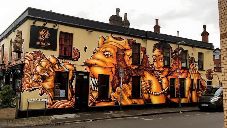 A vividly painted building exterior features a large, intricate mural with orange and yellow hues. The artwork depicts mythological creatures and a regal figure adorned with jewelry. The building has a sign that reads Golden Lion and is located on a corner street.