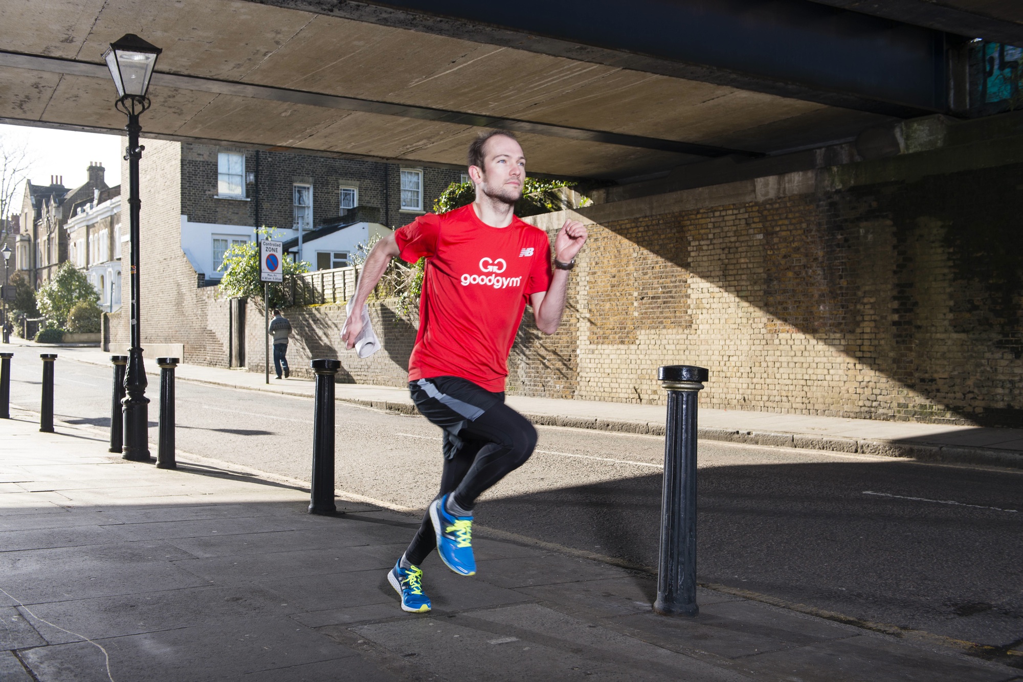 A man in a red GoodGym shirt is running outdoors during the day. He's on a sidewalk under an overpass. The background features a lamp post, street sign, and residential buildings. He's mid-stride, holding a white object in his right hand.