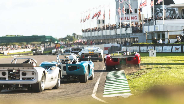 Classic race cars are competing on a track, with one red car slightly off the road onto the grassy shoulder. Spectators watch from the grandstand, while the track is lined with British flags fluttering in the wind on a sunny day.