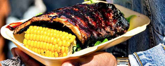 A person holding a cardboard tray with a serving of barbecue ribs coated in sauce, accompanied by two half-cobs of grilled corn. A denim sleeve and wristwatch are partially visible, indicating casual, outdoor dining.