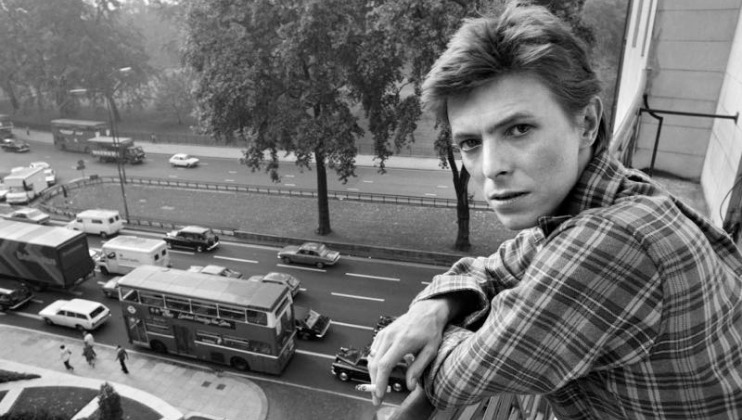 A person with short hair and a plaid shirt leans on a balcony railing overlooking a busy street with cars, buses, and large trees in the background. The person gazes towards the camera with a serious expression.