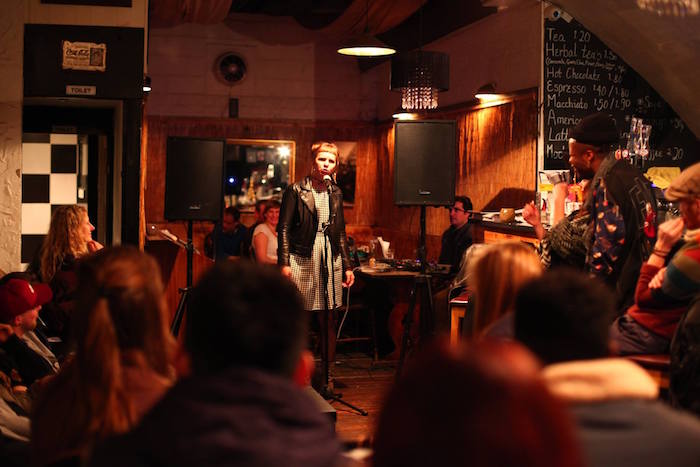 A woman stands at a microphone on a small stage in a cozy, dimly lit coffeehouse. She is speaking or performing to an engaged audience seated around the stage. On the right, a man looks towards her. Menu items and prices are visible on a chalkboard behind them.