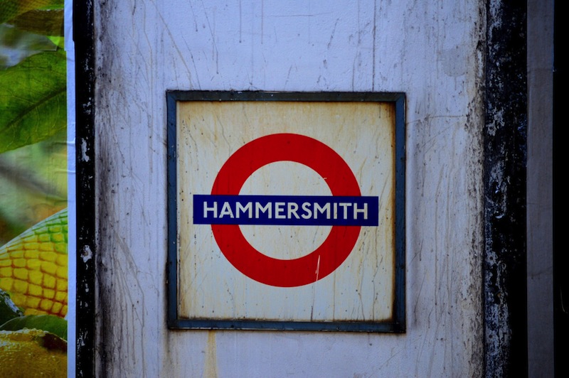 A weathered sign with the London Underground logo, featuring a red circle with a horizontal blue bar across the center. The bar reads HAMMERSMITH in white. The sign is mounted on a wall and shows signs of age and grime.