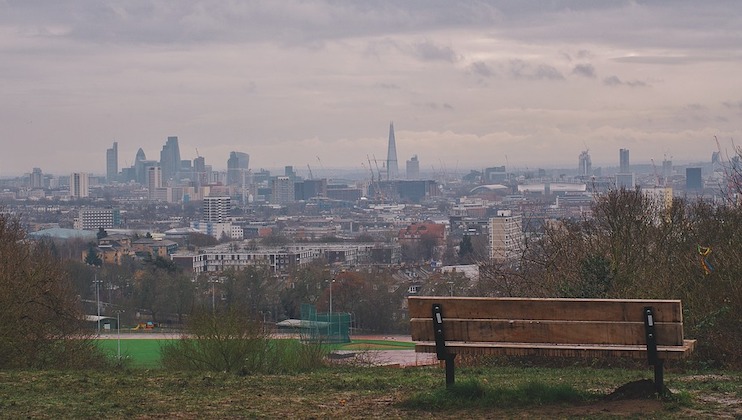 A wooden bench sits in the foreground on a hill overlooking a sprawling cityscape. The skyline, dominated by modern buildings and a prominent skyscraper, stretches under a cloudy sky. Trees and a field are visible closer to the bench.