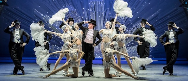 Performers dressed in white, glittering outfits, and top hats are dancing on stage. They are holding large white feathers and striking various poses in front of a sparkling, art deco-style background.