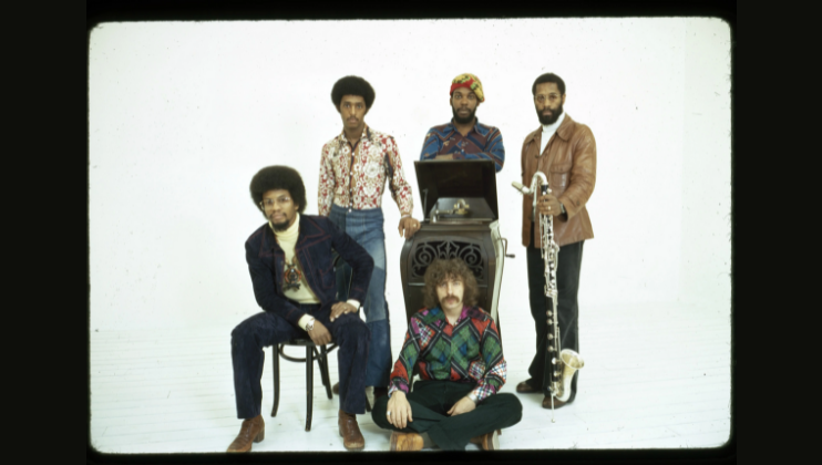A vintage photo of five men posing together. Two men are seated, one on a chair and the other on the floor. The other three stand, one holds a saxophone. They wear colorful, patterned 1970s-style clothing. An ornate music stand is placed in the center.