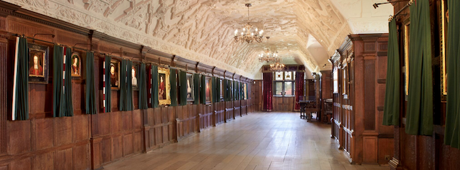A spacious, long corridor with a high, ornately decorated ceiling and wooden paneling on the walls. The corridor is adorned with framed portraits on either side, partially covered by green curtains. Chandeliers hang from the ceiling, illuminating the space warmly.