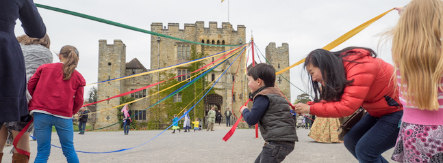 People, including children, are participating in maypole dancing in front of a large historic stone building with battlements. Colorful ribbons are attached to the top of a pole and held by the participants as they weave and dance around it.