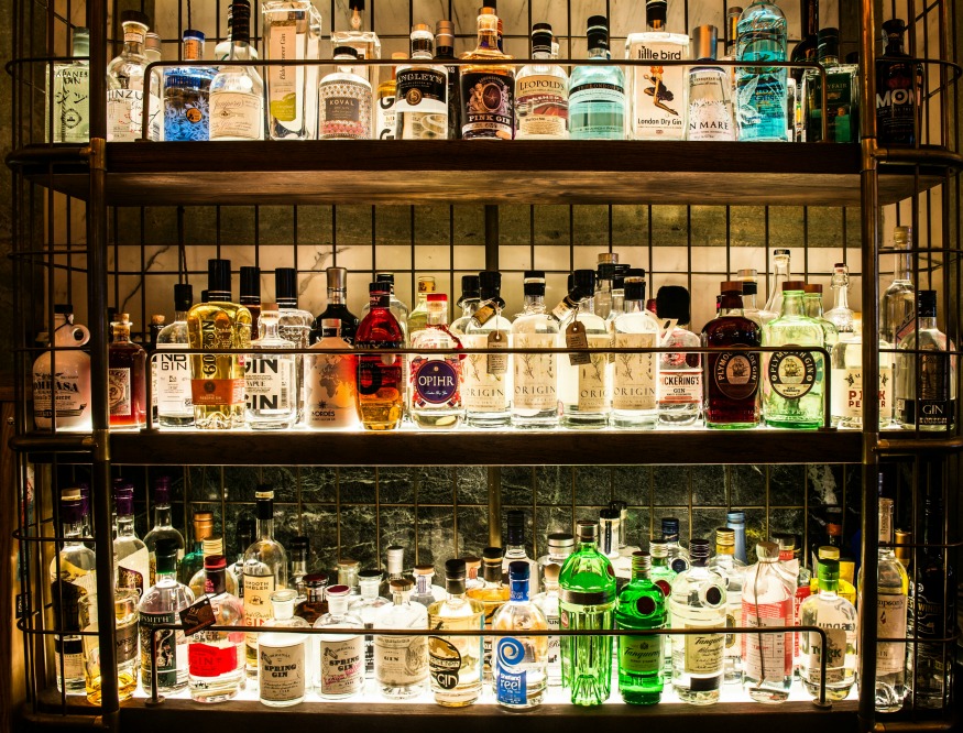 A collection of various liquor bottles is displayed on multiple shelves. Bottles of different shapes, sizes, and colors are arranged neatly. The shelves are backlit, highlighting the variety of labels and brands, and creating a warm, inviting ambiance.
