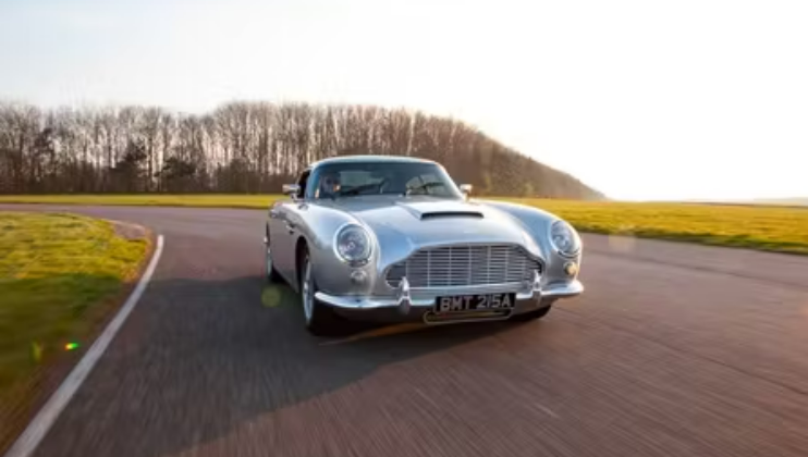 A classic silver car with a sleek design is driving on a curved open road under a clear sky. The license plate reads BMT 216A. A line of trees in the background suggests it is autumn or winter, as the trees are without leaves.