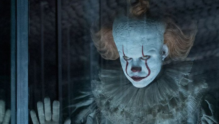 A menacing clown with white face paint, red nose, exaggerated red lips, and wild orange hair, wearing a ruffled collar, presses its hands against a glass barrier. The background is dark and foreboding, adding to the eerie atmosphere.