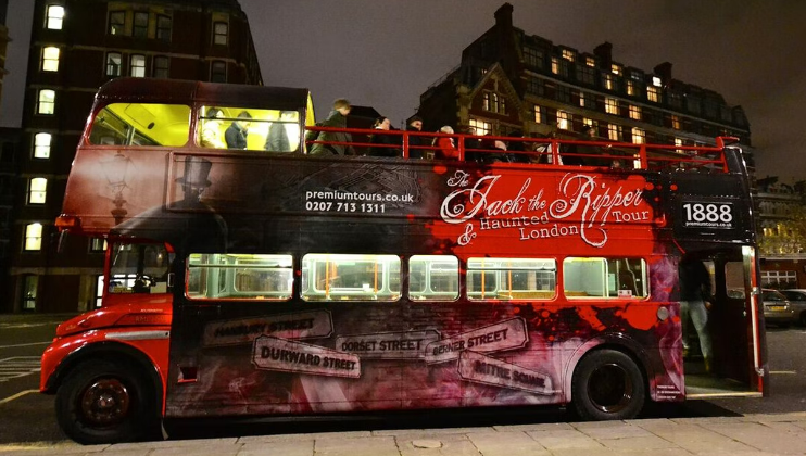 A double-decker bus designed for a Jack the Ripper Haunted London Tour is parked on a city street at night. The bus is decorated with eerie graphics and historical references to Jack the Ripper's infamous sites. Passengers are seated on the partially open upper deck.