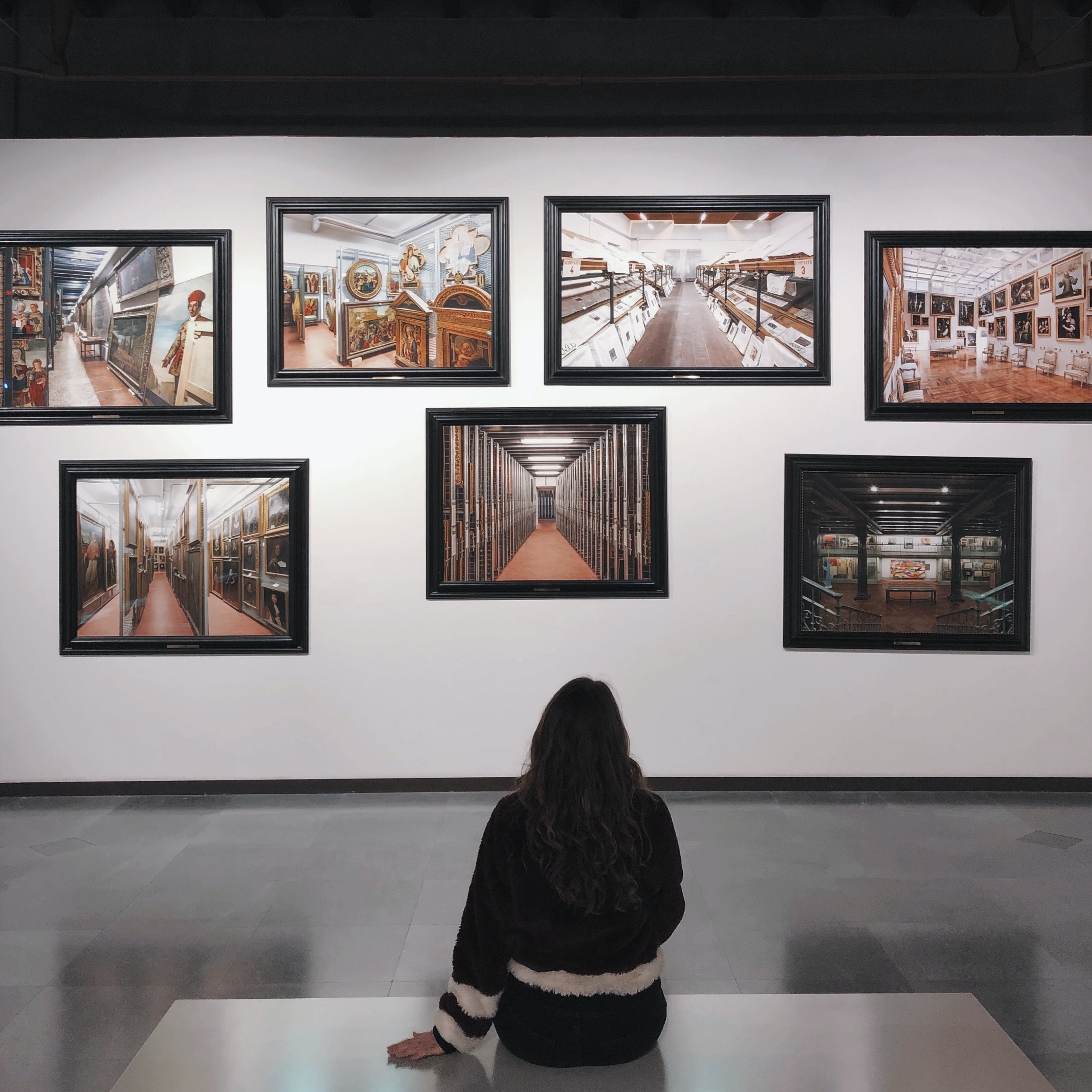 Lady standing on a white bench inside a gallery looking at 9 paintings of varying sizes on a grey wall