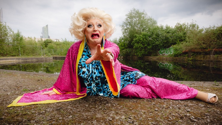 A person with curled blonde hair dressed in a vibrant pink and yellow outfit with a blue patterned top is sitting on the ground near a body of water, reaching out with an expressive face. Bushes and a building can be seen in the background.