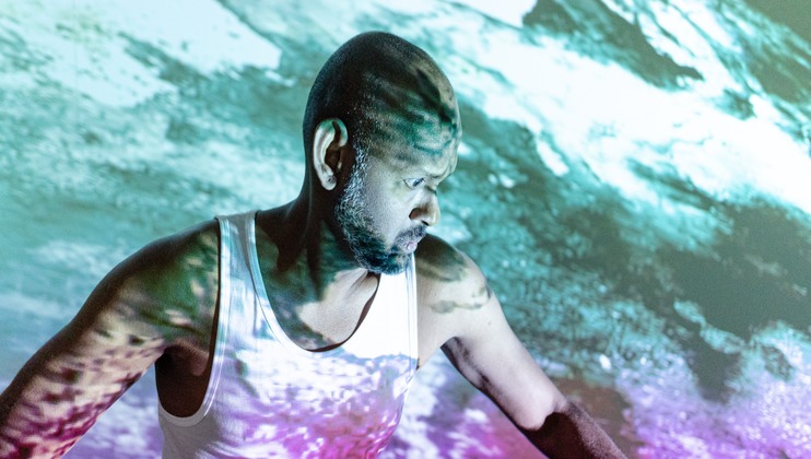 A man with a shaved head and beard is wearing a white tank top. He is bathed in colorful light projections, predominantly green and purple, creating an abstract, dynamic background. He looks to the side with a serious expression.
