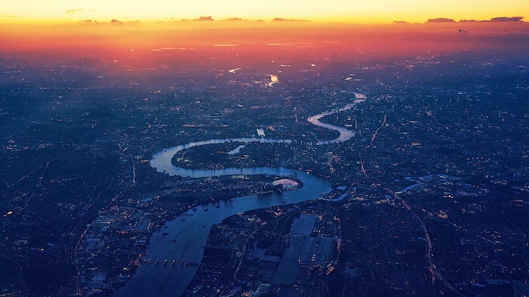 Aerial view of a city at sunset with a winding river running through it. The sky is filled with vibrant hues of orange and purple, while the lights of the city are starting to illuminate the landscape below. The scene evokes a sense of tranquility and beauty.