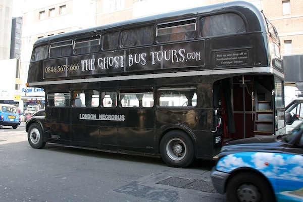 A black double-decker bus labeled London Necrobus offers The Ghost Bus Tours.com with a phone number. The bus is parked on a city street with buildings in the background and other vehicles nearby. The bus has an open door revealing a glimpse of red interior stairs.