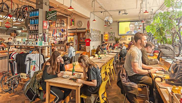 A bustling café with a cozy and eclectic vibe. Patrons sit at wooden tables, some working on laptops while others chat and enjoy coffee. The walls are adorned with cycling gear, and there's a mix of plants, books, and art, creating a lively atmosphere.