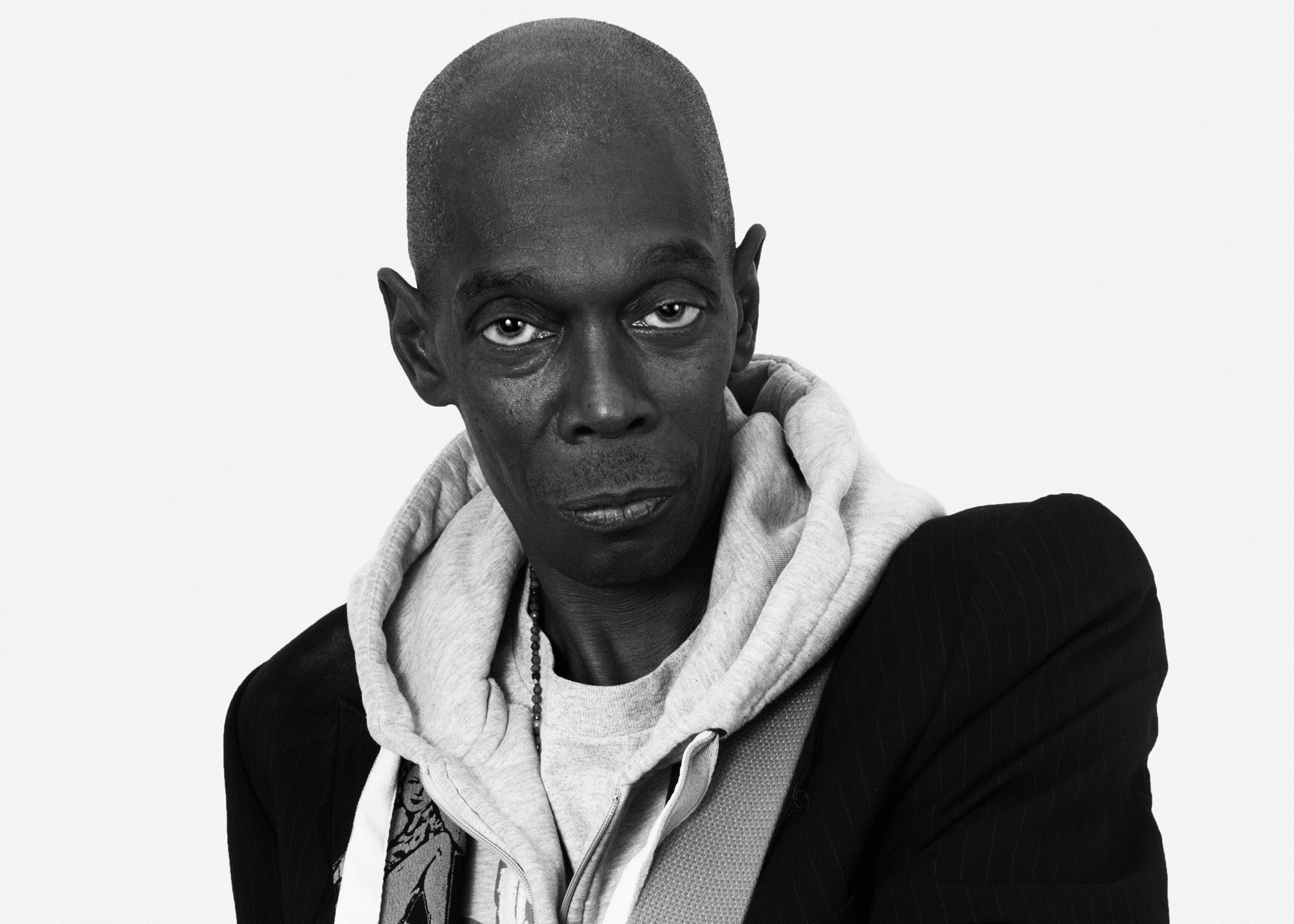 A black and white portrait of a person looking directly at the camera. They have a shaved head and wear a layered outfit consisting of a hoodie and a jacket. Their expression is neutral. The background is plain, emphasizing the subject's features and clothing.