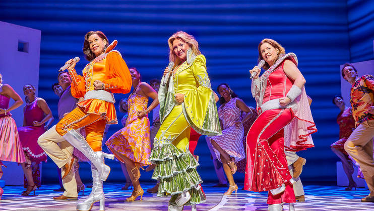 Three performers are on stage wearing brightly colored costumes. The person on the left is in an orange outfit, the person in the center is in green, and the person on the right is in red. Each holding a microphone, they sing and dance energetically with a group of dancers in the background.