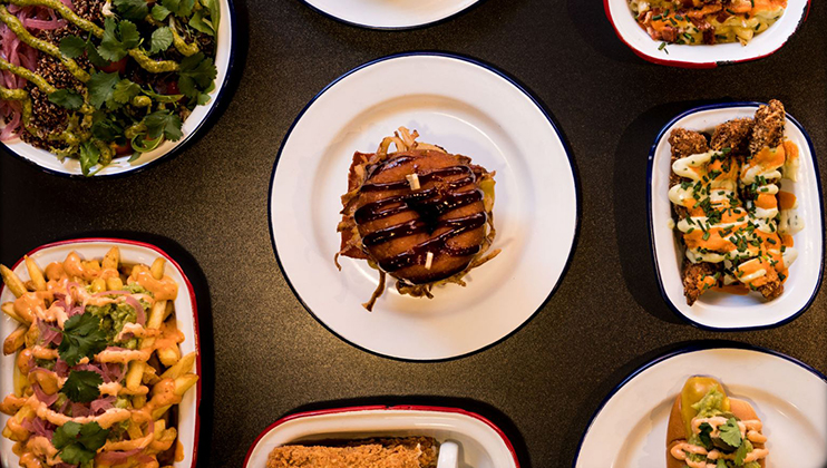 A top-down view of various plated dishes on a dark table. Central is a main dish with a burger and fries. Surrounding are sides including loaded fries, salad, fried chicken, and a baked dish topped with sauce and herbs, all presented in white and red-rimmed plates.