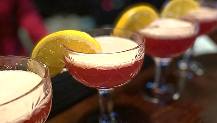 A close-up shot of four cocktails served in glass coupe glasses, each garnished with a slice of lemon on the rim. The beverages have a pinkish hue with a frothy top, arranged in a line on a dark wooden bar. The background shows a blurred figure.
