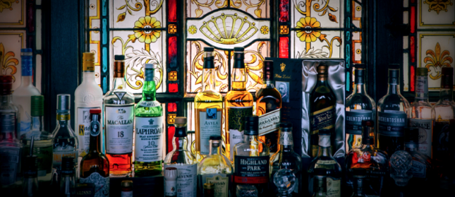 A variety of whiskey bottles are displayed on a wooden surface in front of an ornate, stained glass window with intricate floral and geometric designs. The bottles include several well-known brands, creating a colorful and varied collection.