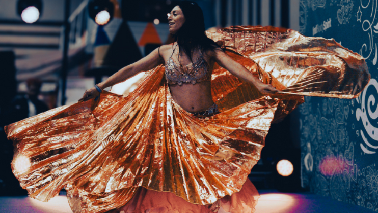 A person performing a dance with golden shimmering wings and a decorated top, creating a majestic, flowing motion. The background is festive, with colorful lights and decorations, suggesting the setting is a lively event or celebration.
