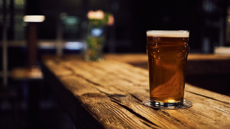 A tall glass of amber beer sits on a wooden bar counter. The beer has a frothy head, and the glass is placed on a coaster. The background is dimly lit, providing a cozy and rustic atmosphere, with blurred lights and objects visible.