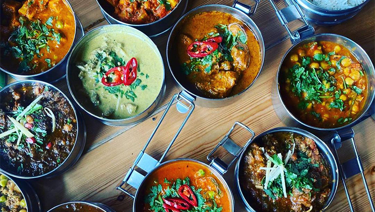 Various bowls of colorful Indian curries and dishes are arranged on a wooden table. The dishes include garnishes such as red chili slices and fresh herbs, presenting a vibrant array of textures and flavors.
