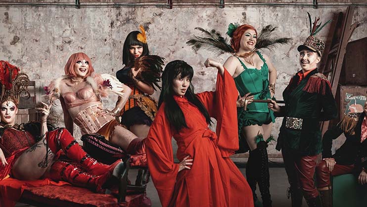 A group of six people dressed in elaborate, colorful burlesque or cabaret-inspired costumes pose in front of an aged, industrial wall. They wear outfits featuring feathers, corsets, and vibrant accessories, with lively expressions and confident stances.