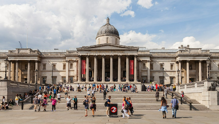 The image shows the National Gallery in London, a neoclassical building with a central dome and columned portico. Visitors are gathered on the steps and plaza in front of the gallery. The sky is partly cloudy, and the area is bustling with activity.