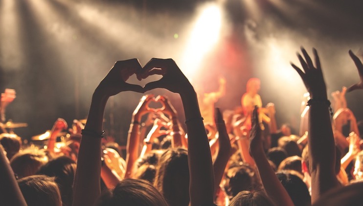 A crowd at a concert is raising their hands, with some people forming heart shapes with their fingers. The stage lights in the background create a vibrant and energetic atmosphere. The scene captures the excitement and unity of the audience.