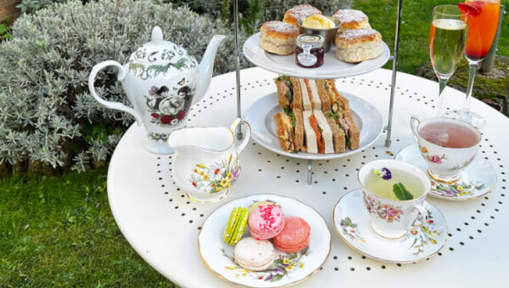An afternoon tea setup on a white table in a garden. The table features a floral teapot, a milk jug, a tiered tray with scones, sandwiches, and macarons, and floral teacups filled with beverages, including green tea, pink tea, and champagne with fruit and flowers.