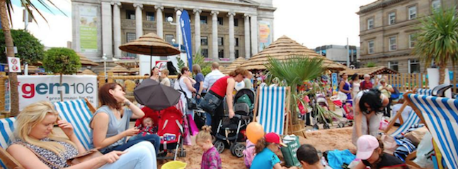 A busy urban beach event in front of a historic building, with people relaxing on deck chairs, children playing in the sand, and strollers scattered around. Palm trees, beach huts, and a gem 106 banner add to the festive atmosphere.